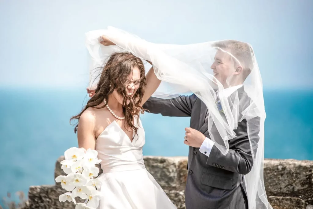 Charlotte Davies Photography - St. Andrews Wedding Photographer. A bride and groom stand outdoors by the sea. The groom is playfully adjusting the bride's veil while she holds a bouquet of white flowers.