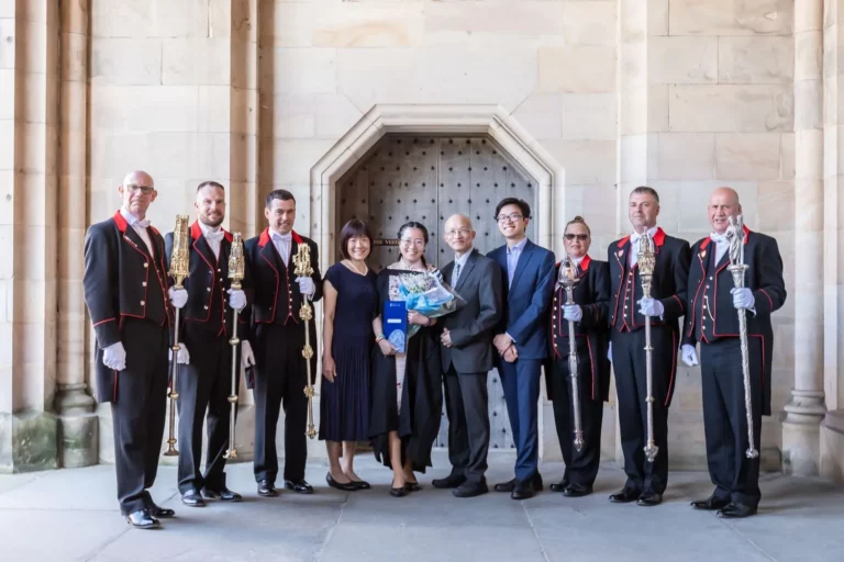 Charlotte Davies Photography - St. Andrews Wedding Photographer. A group of eight people poses, six in formal uniforms holding staffs, and two in suits, standing in front of a stone wall with an arched doorway.