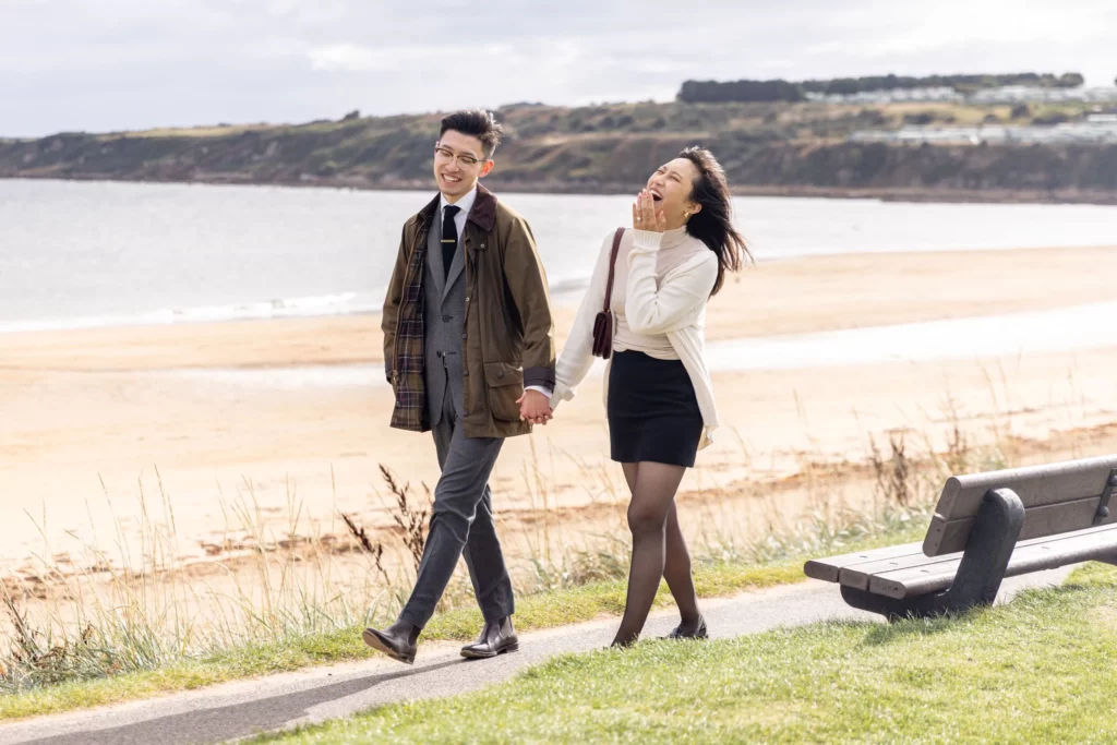 Charlotte Davies Photography - St. Andrews Wedding Photographer. A couple walks hand in hand along a seaside path, with the beach and ocean in the background. They appear to be enjoying a leisurely stroll on a windy day.