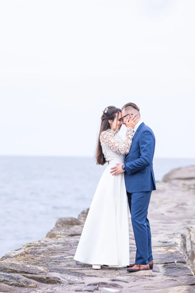 Charlotte Davies Photography - St. Andrews Wedding Photographer. A couple embraces on a rocky shoreline. The woman wears a white dress, and the man is in a blue suit. The ocean is in the background under a cloudy sky.