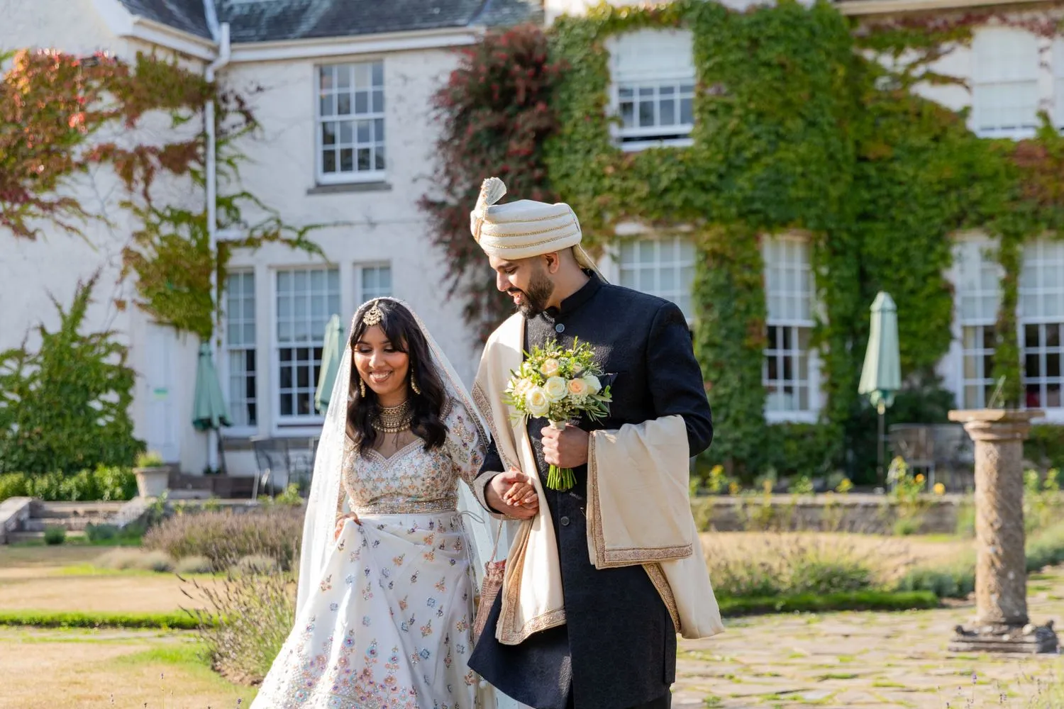 Charlotte Davies Photography - St. Andrews Wedding Photographer. A couple in traditional attire walks outdoors, holding hands. The woman wears a floral lehenga, and the man wears a sherwani with a turban. They are in front of a vine-covered building.