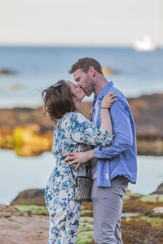 Charlotte Davies Photography - St. Andrews Wedding Photographer. A couple stands near beach rocks, embracing and kissing. The man wears a blue shirt and gray pants, while the woman wears a floral dress. The ocean is visible in the background.