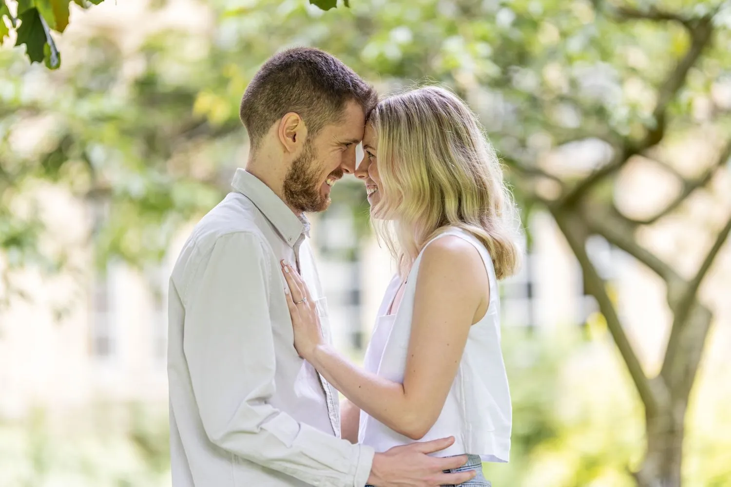 Charlotte Davies Photography - St. Andrews Wedding Photographer. A man and woman stand closely, smiling, with their foreheads touching. They are outdoors with blurred trees in the background.