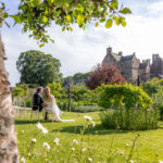 Newlyweds seated in a garden beside a historic stone castle