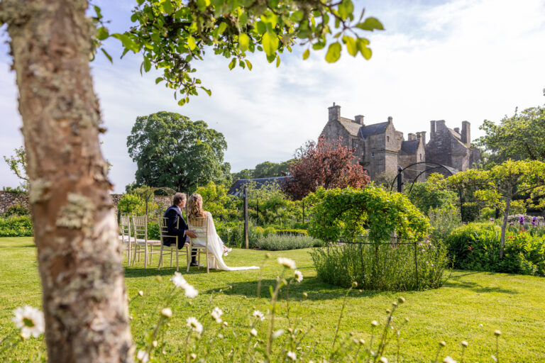 Newlyweds seated in a garden beside a historic stone castle