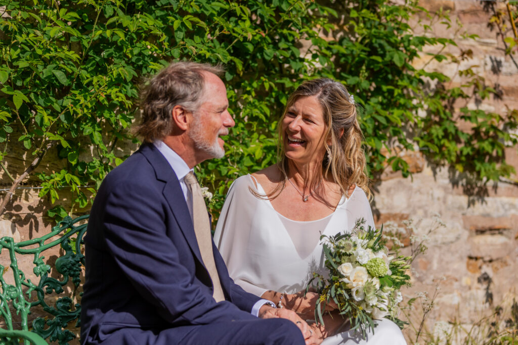 Laughing newlyweds seated beside a leafy stone wall in the walled garden at Kellie Castle, Fife.