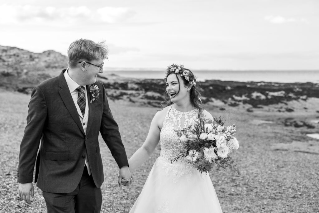 Laughing newlyweds hold hands on a rocky beach
